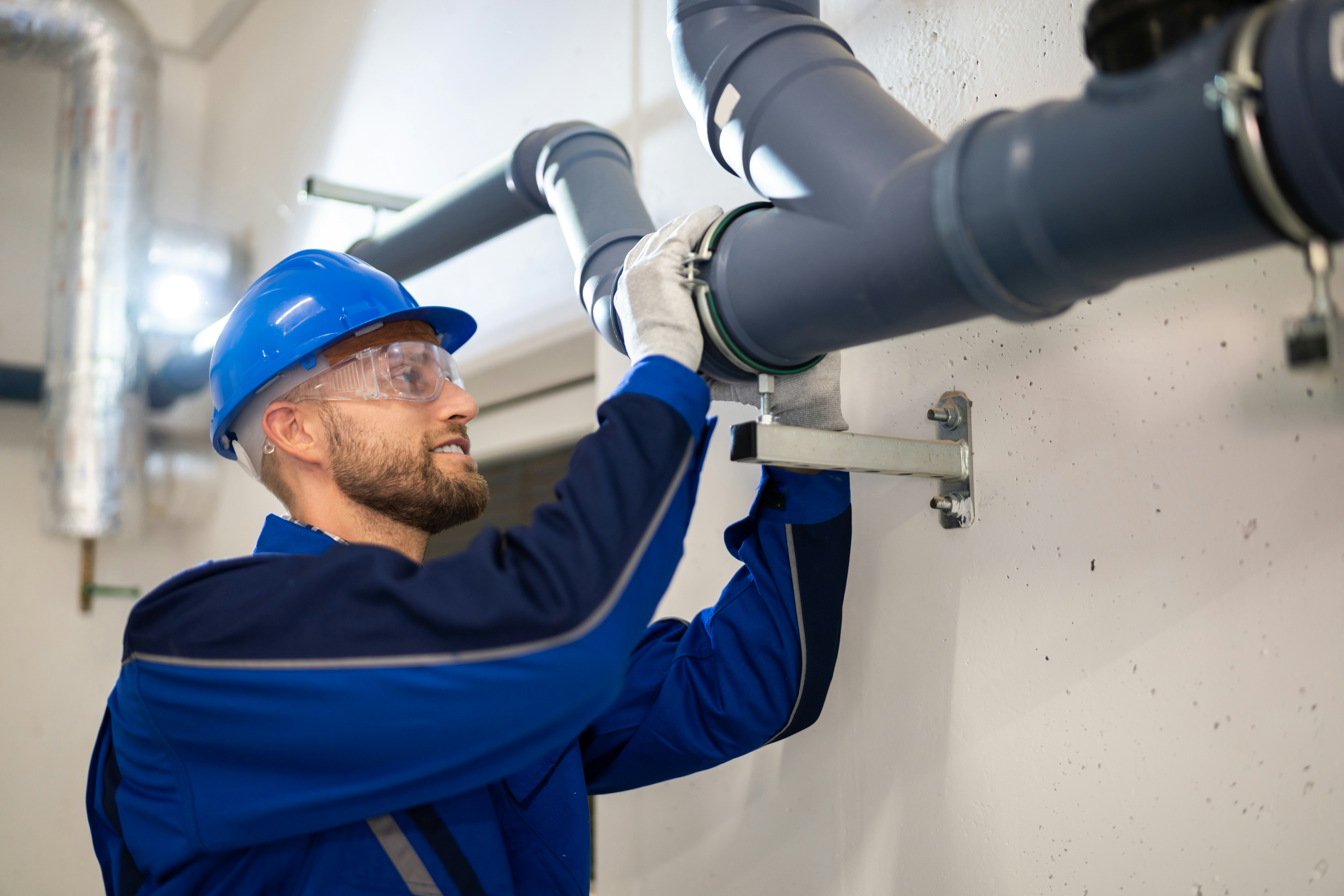 Construction worker in blue uniform and hard hat inspecting plumbing pipes.